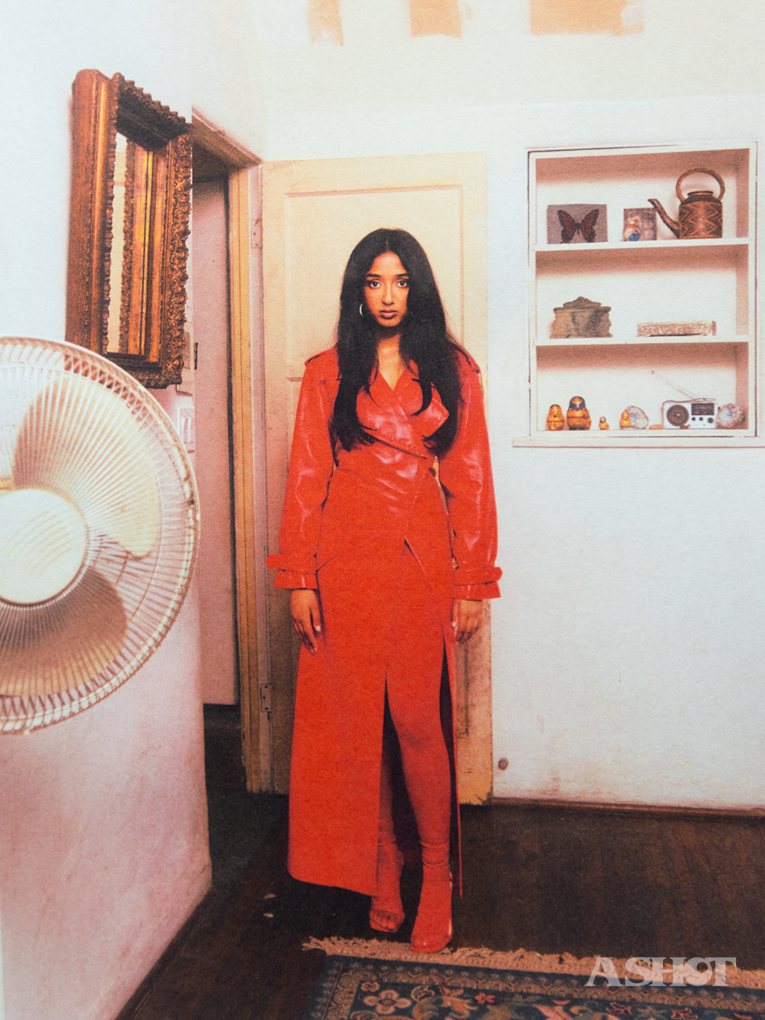 Maitreyi Ramakrishnan standing in a red trench coat facing the camera in a run down room